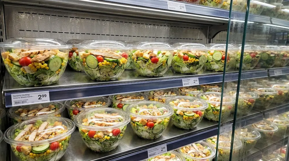 32 oz clear PET salad bowls displayed in a supermarket chilled shelf with chicken salad, vegetables, corn, tomatoes, and cucumber