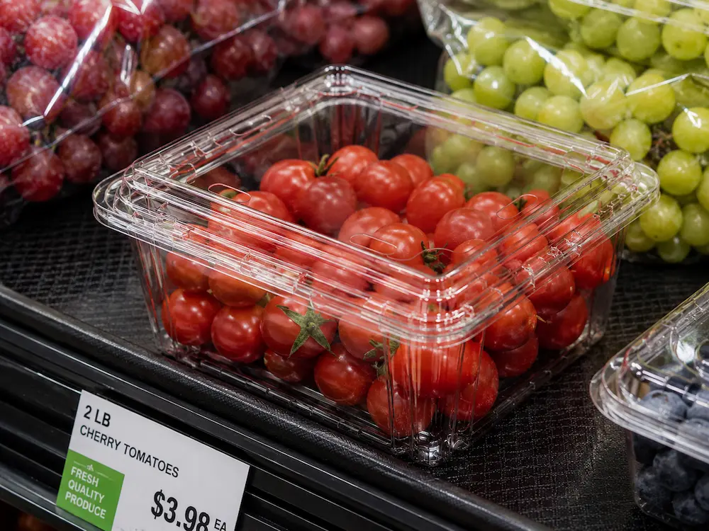 Clear PET cherry tomato clamshell tray displayed with grapes and fresh produce in a supermarket case.