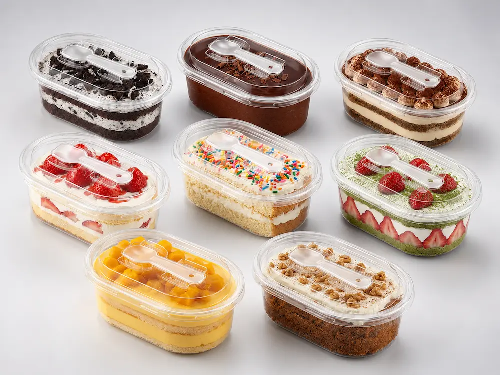 Clear PET/RPET oval dessert containers with spoon in assorted cake, fruit, chocolate, matcha, mango, and tiramisu dessert styles on a light cool gray studio background