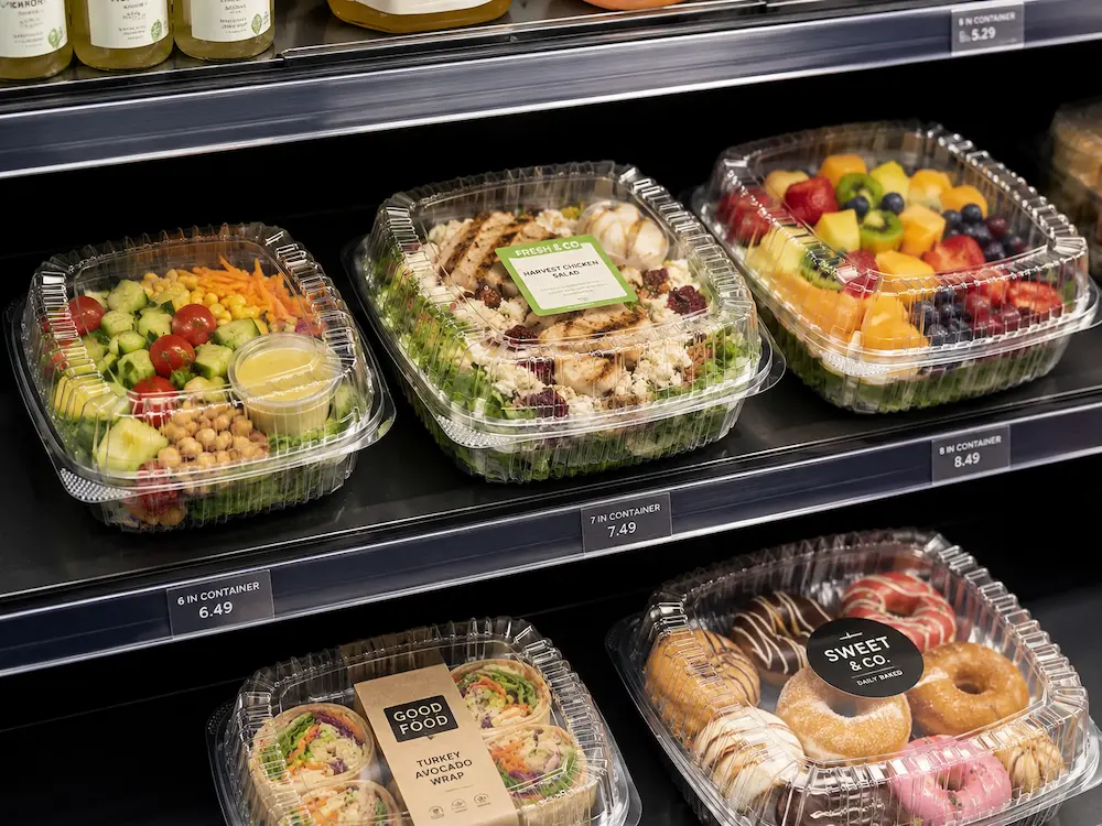PET hinged deli containers used in a retail refrigerated display for salads, fruit, wraps, and bakery items.