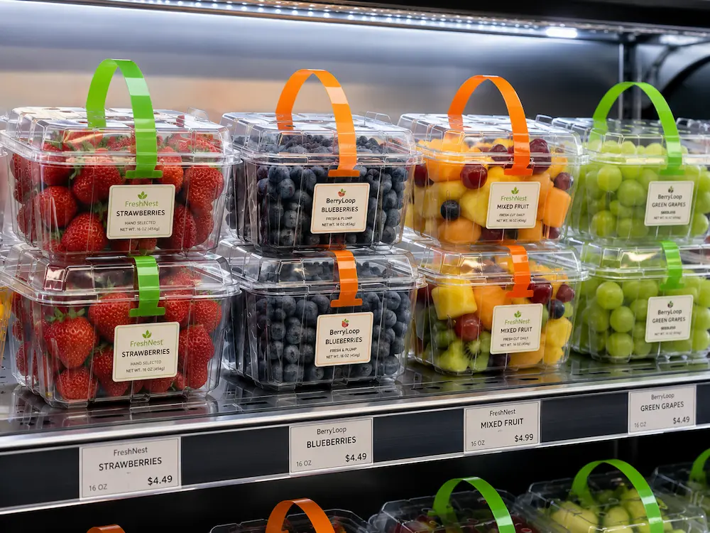 Clear fruit containers in refrigerated grab-and-go display with labels and stacked PET packaging