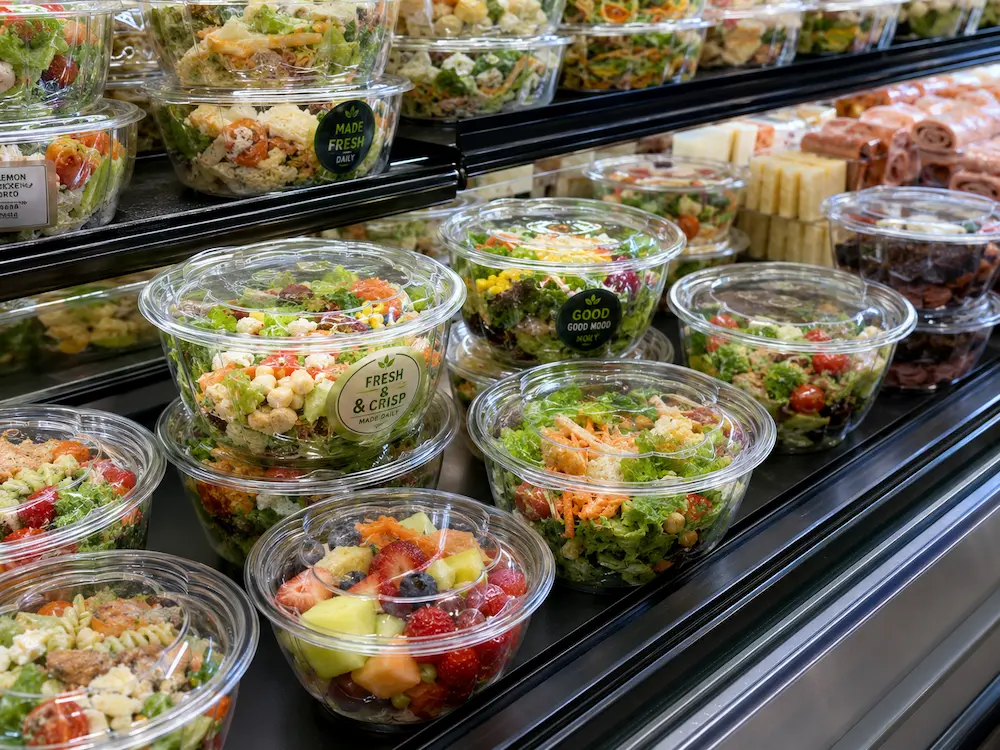 Round PET deli containers displayed in a supermarket chilled shelf with salads, fruit, and prepared foods
