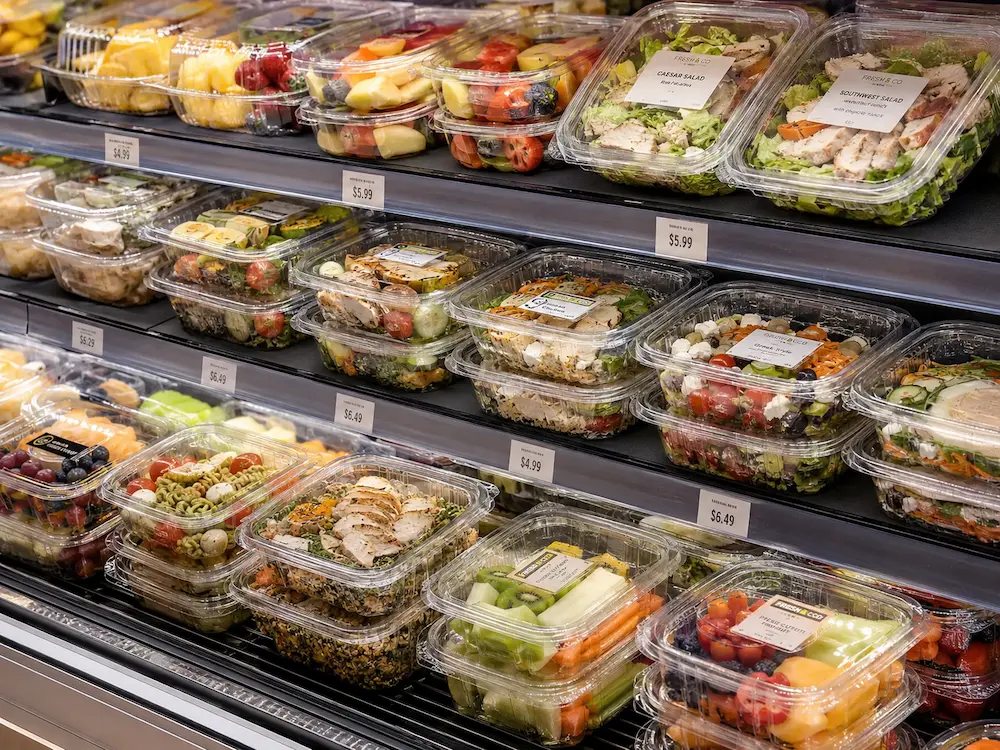 Clear PET food containers displayed in a supermarket refrigerated case with salads, fruit, and ready-to-eat meals