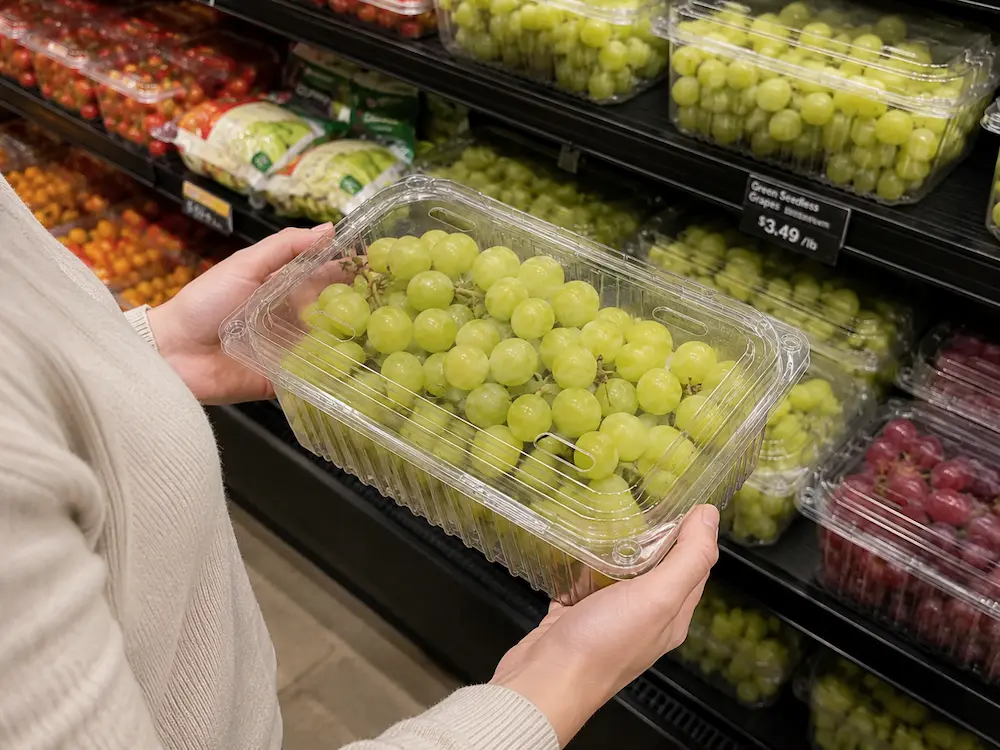 Shopper holding a clear PET vented grape clamshell in a supermarket fresh produce aisle