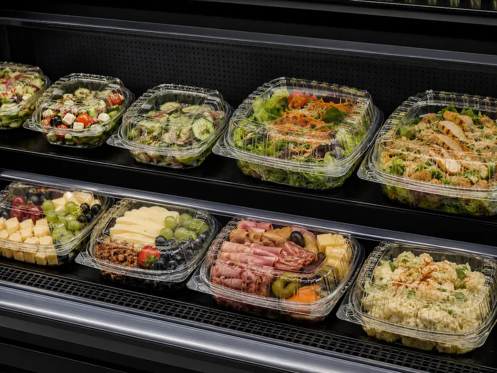 Clear PET deli clamshell containers displayed with salads, fruit, pasta, and deli foods in a refrigerated retail case