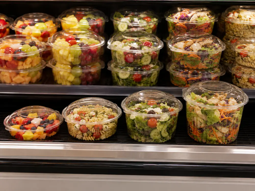 Clear round deli containers displayed with salad and fruit in refrigerated retail shelf
